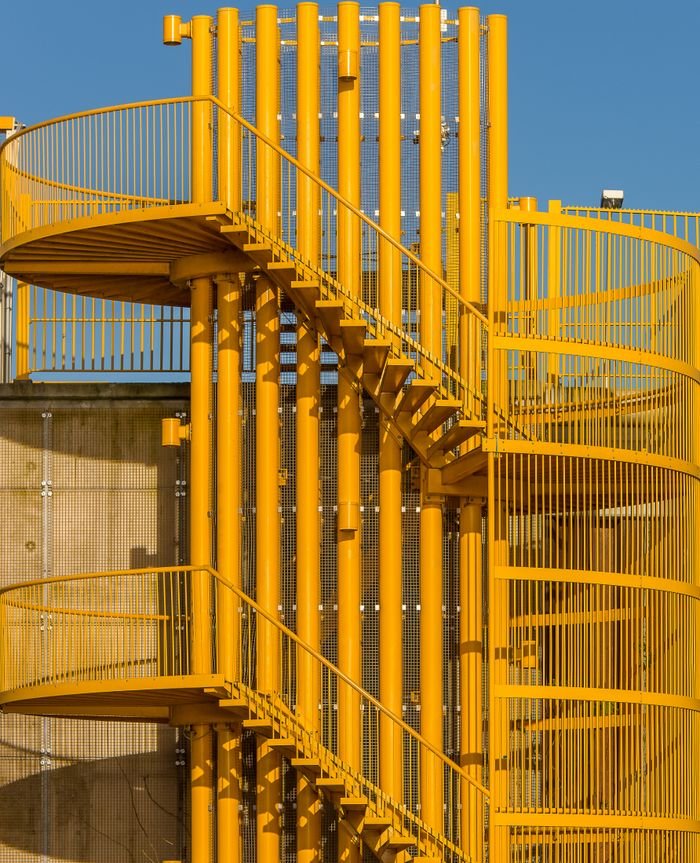 Bright yellow metal spiral staircase with architectural design elements, set against a clear blue sky.